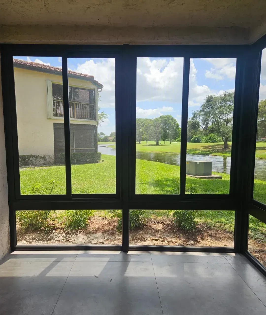 Dark-framed glass patio enclosure with a tile floor, looking out at a lake and green lawn